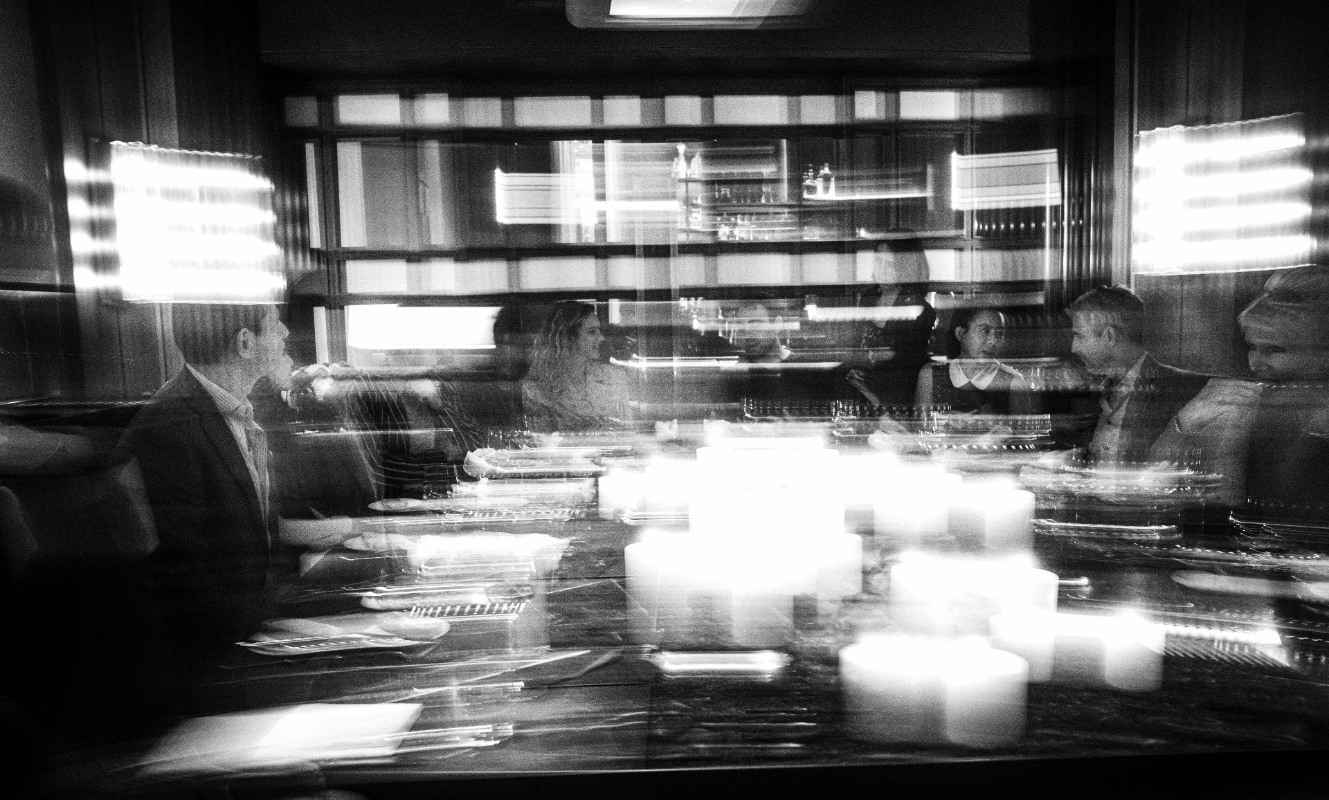 Long-exposure black-and-white photograph of guests around a candlelit dinner table